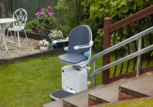 Εξωτερικό αναβατόριο σκάλας με γκρι κάθισμα, τοποθετημένο σε σκάλα κήπου. Το αναβατόριο βρίσκεται στη βάση της σκάλας, δίπλα σε γκαζόν και λουλούδια.