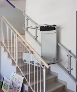 Αναβατόριο πλατφόρμα για αμαξίδιο, εγκατεστημένο σε εσωτερική σκάλα με λευκά κάγκελα. Η πλατφόρμα είναι διπλωμένη και παρκαρισμένη στη μέση της διαδρομής.