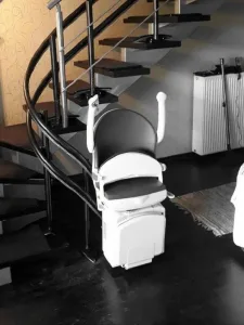 Αναβατόριο καρέκλα σκάλας με το κάθισμα διπλωμένο, σταθμευμένο στη βάση μιας μοντέρνας καμπυλωτής σκάλας με σκούρα πατήματα.