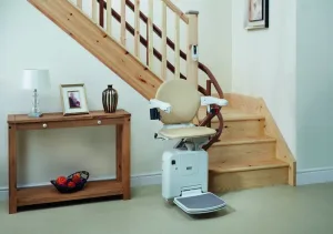 Αναβατόριο σκάλας με μπεζ κάθισμα, σταθμευμένο στη βάση μιας ξύλινης σκάλας, δίπλα σε ένα ξύλινο τραπέζι κονσόλα.